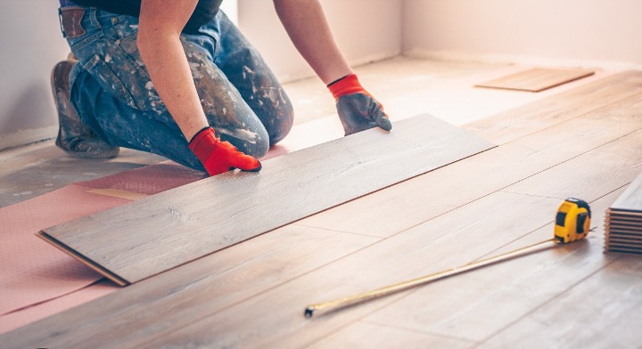 Pracownik w czerwonych rękawicach montuje panel podłogowy na różowym podkładzie, obok leży żółta miarka.