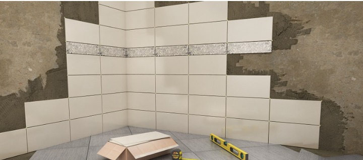 Proces układania beżowych płytek ściennych w rogu pomieszczenia, z dekoracyjnym pasem płytek z motywem roślinnym, widoczne narzędzia: poziomica i miarka.