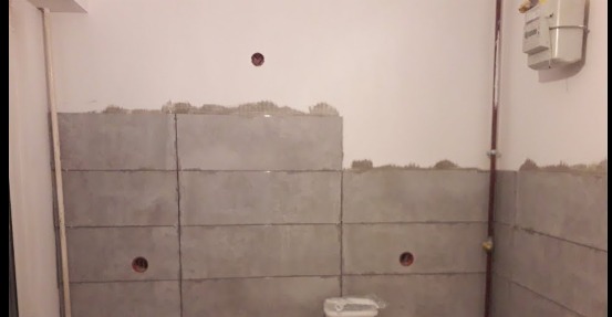 Ściana w trakcie wykańczania z szarymi panelami, widoczne puszki elektryczne i pionowa rura z gazomierzem w rogu pomieszczenia.