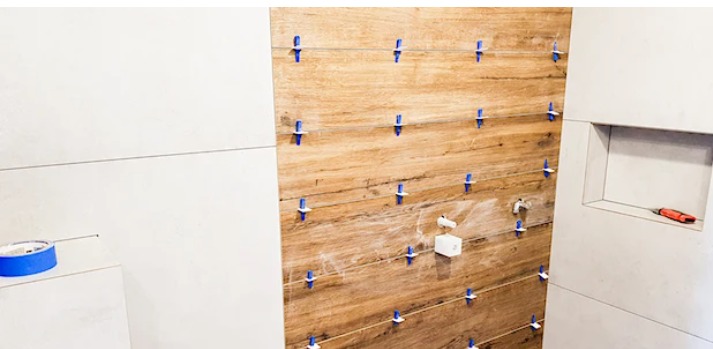 Ściana łazienki w trakcie wykańczania z drewnopodobnymi płytkami, użyto systemu poziomowania z niebieskimi klipsami, widoczna instalacja wodna i wnęka z pomarańczowym śrubokrętem w tle.