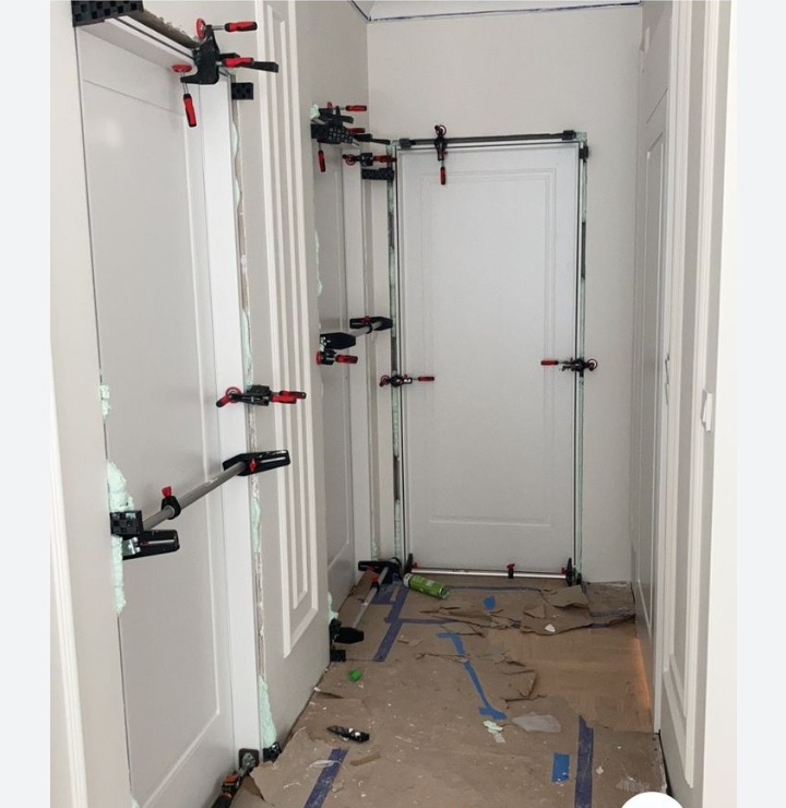 Montaż ościeżnic drzwiowych w korytarzu, widoczne pianka montażowa i specjalistyczne rozpórki mocujące, podłoga zabezpieczona kartonem.
