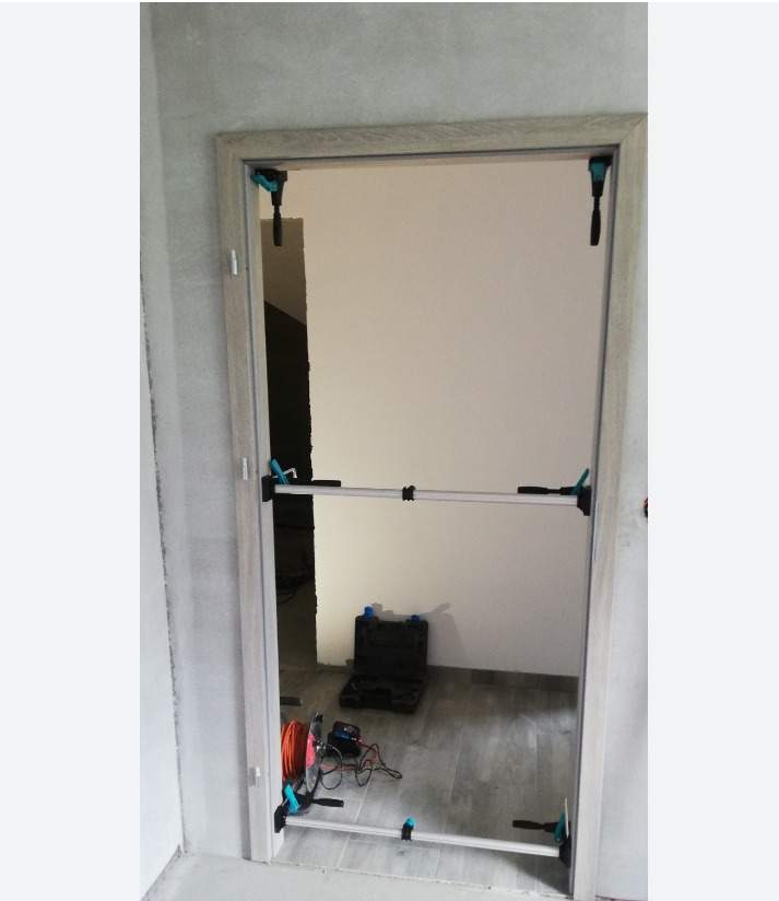 Montaż ościeżnicy drzwiowej z użyciem rozpórki teleskopowej Wolfcraft, widoczny przewód elektryczny i skrzynka narzędziowa w tle.