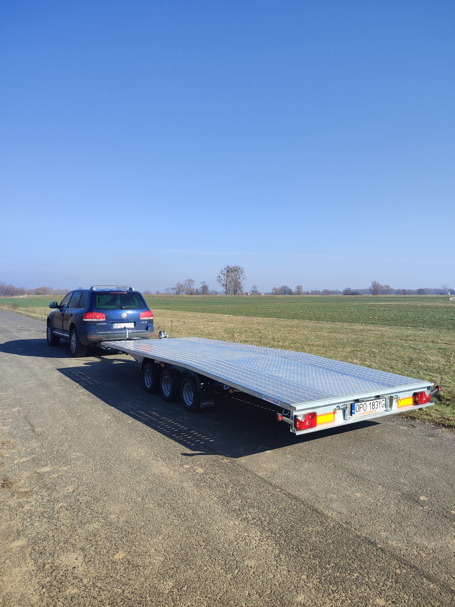 Niebieski samochód osobowy marki Volkswagen Touareg ciągnie srebrną, czterokołową przyczepę lawetę na asfaltowej drodze, obok pola, w słoneczny dzień.