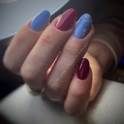 Dłoń z pomalowanymi paznokciami: dwa w kolorze bordowym, dwa w kolorze błękitnym z drobnymi, białymi kropeczkami, prezentowane w zbliżeniu.