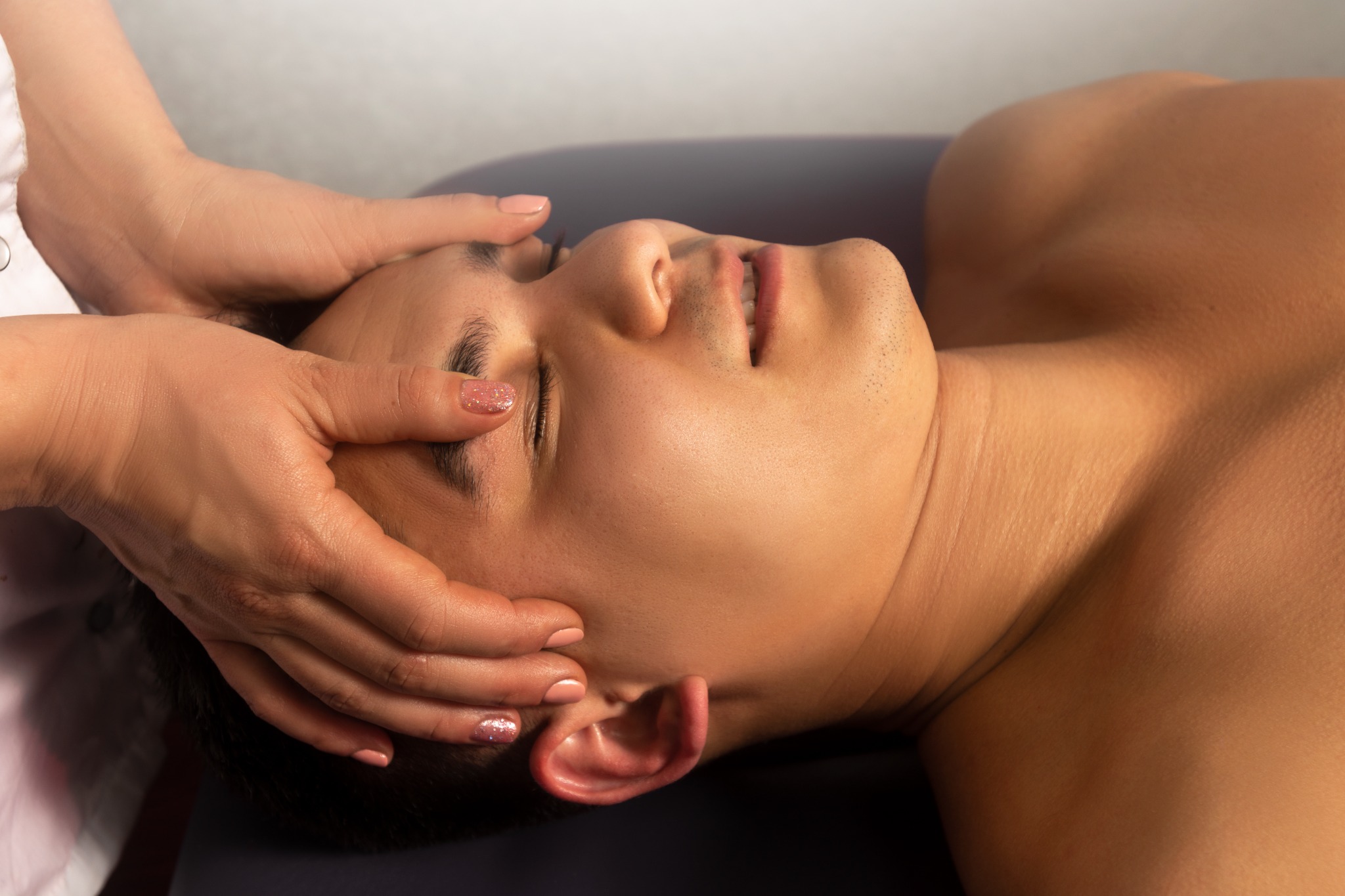 Mężczyzna z zamkniętymi oczami podczas delikatnego masażu głowy, dłonie masażystki z jasnym lakierem na paznokciach, widoczne delikatne zmarszczki na czole, oświetlenie podkreśla kontury twarzy.