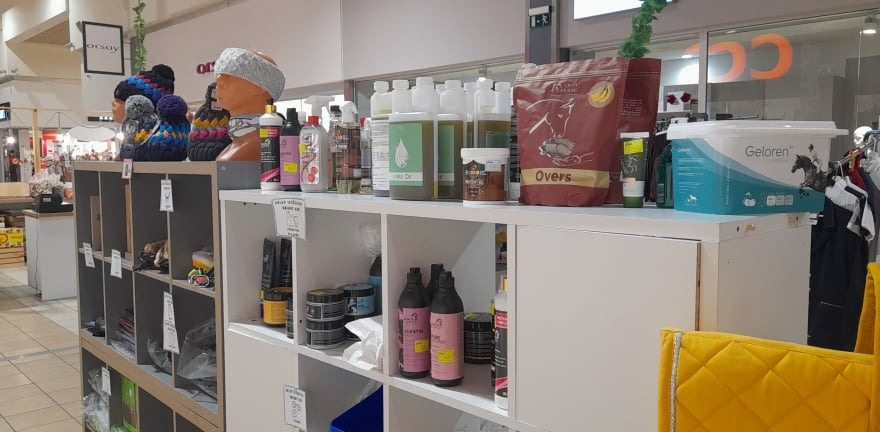 Półki sklepowe z produktami dla koni: suplementy, oleje, szampony i akcesoria jeździeckie, widoczne manekiny z czapkami i opaskami.