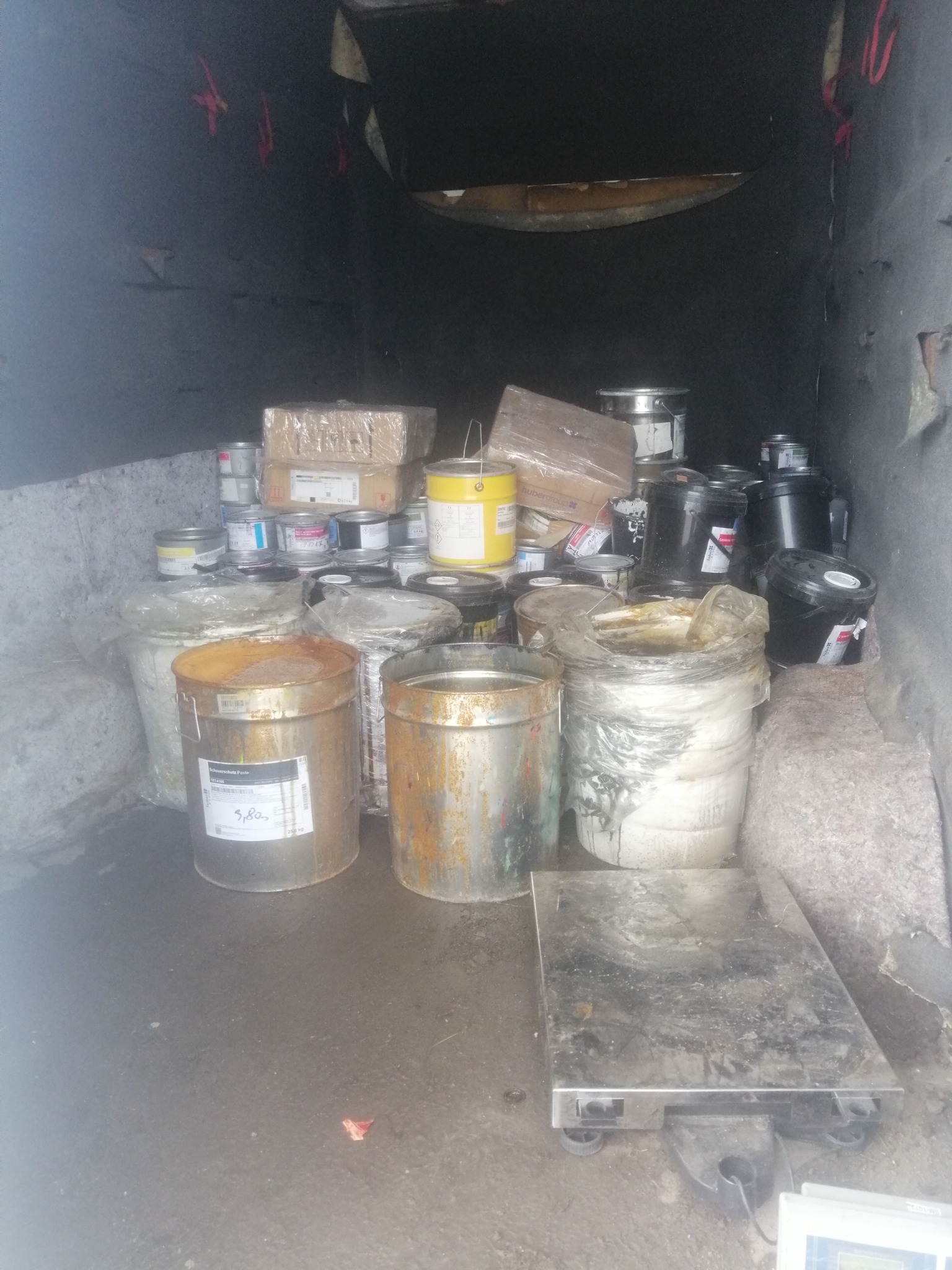 Wnętrze brudnej paki samochodu dostawczego wypełnione różnymi pojemnikami z farbami, lakierami i innymi substancjami chemicznymi, obok stoi waga platformowa.