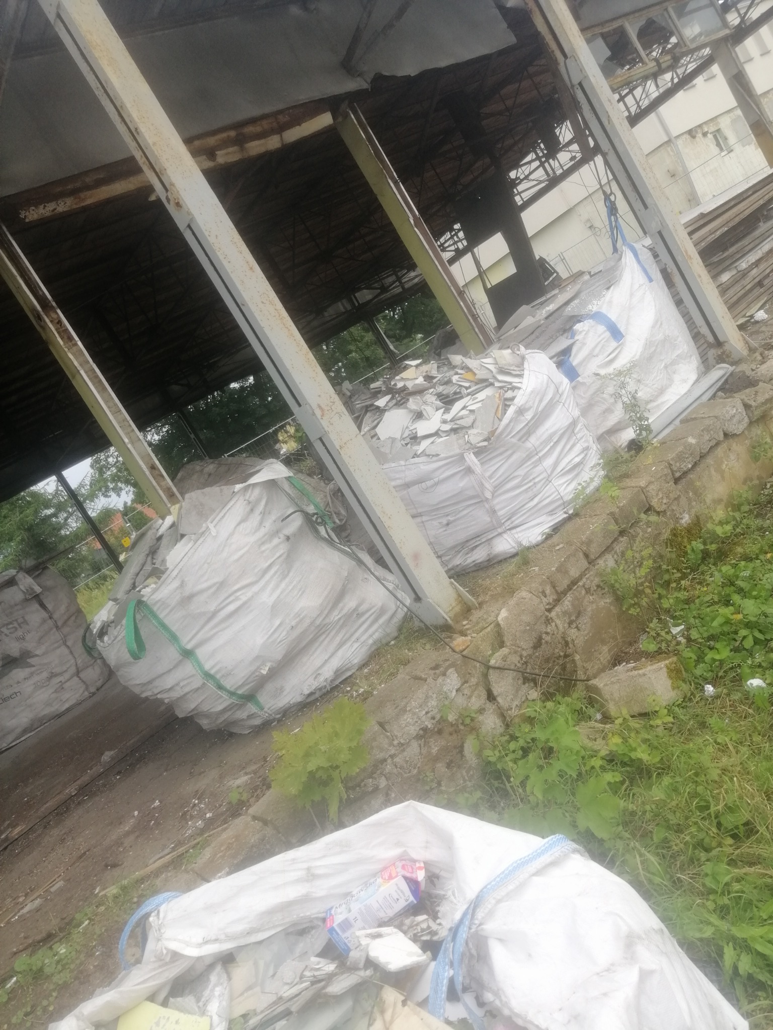 Worki typu Big Bag wypełnione gruzem budowlanym, składowane pod zadaszeniem konstrukcji stalowej, widoczne elementy konstrukcyjne budynku w tle.