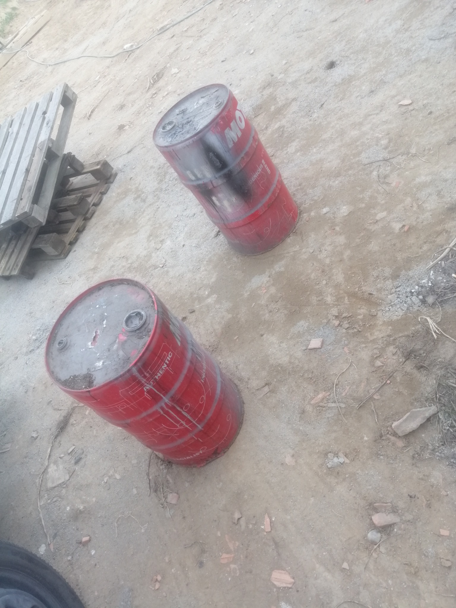 Dwa czerwone, metalowe beczki leżące na ziemi z gruzem i fragmentami cegieł, w tle paleta drewniana.