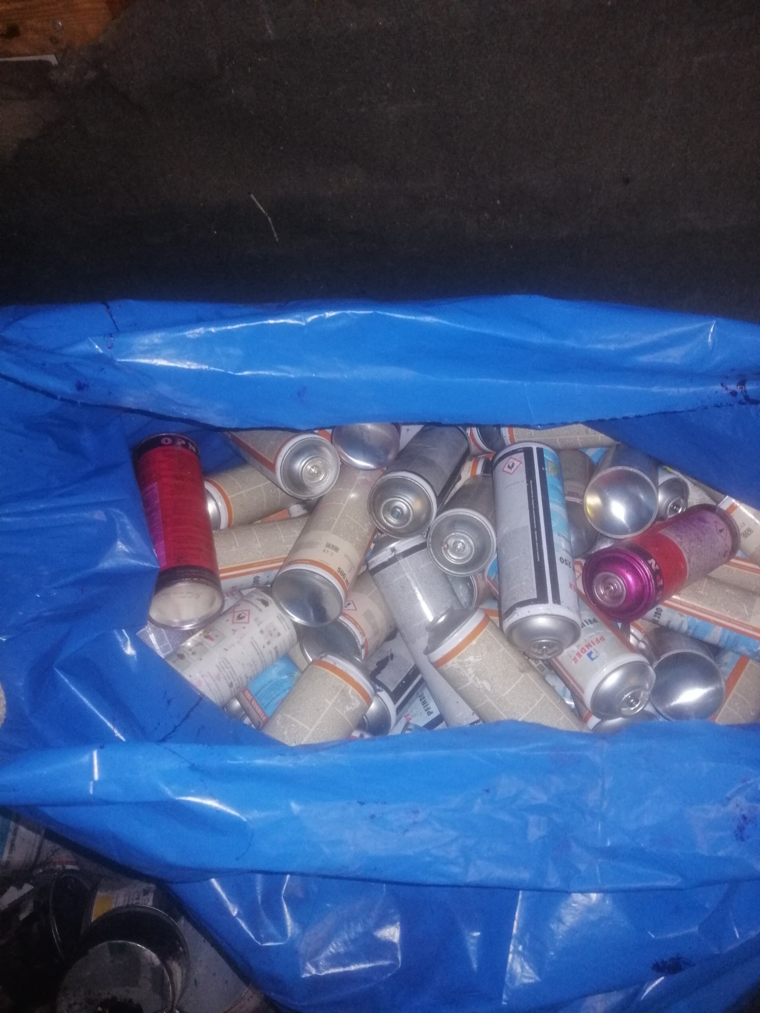 Niebieski worek wypełniony zużytymi puszkami po farbach w sprayu różnych kolorów i rozmiarów, widoczne etykiety i oznaczenia ostrzegawcze.
