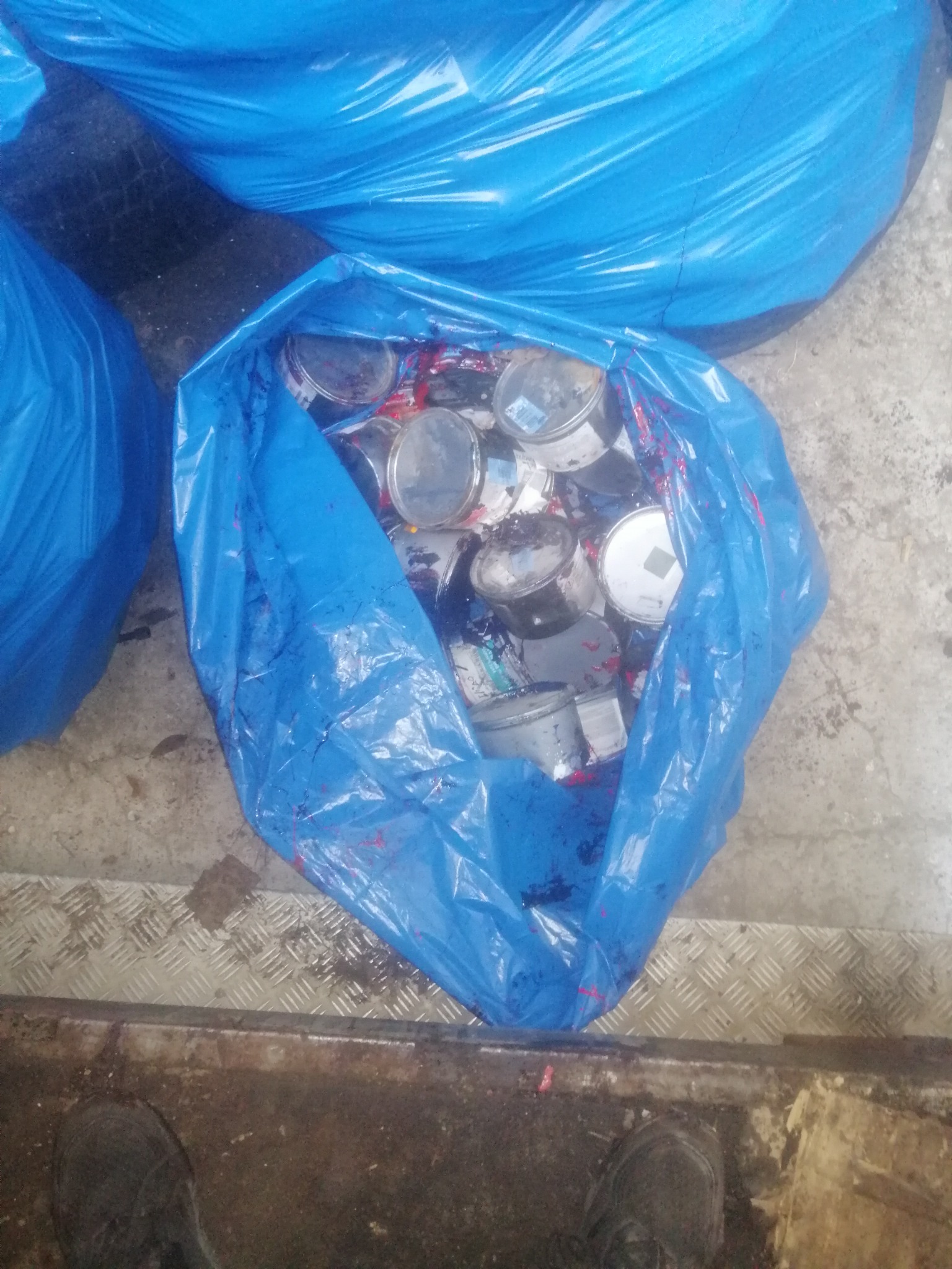 Niebieski worek wypełniony pustymi, metalowymi puszkami po farbach, stojący na metalowej powierzchni z ryflowanym wzorem, widoczne ślady zaschniętej farby na worku.