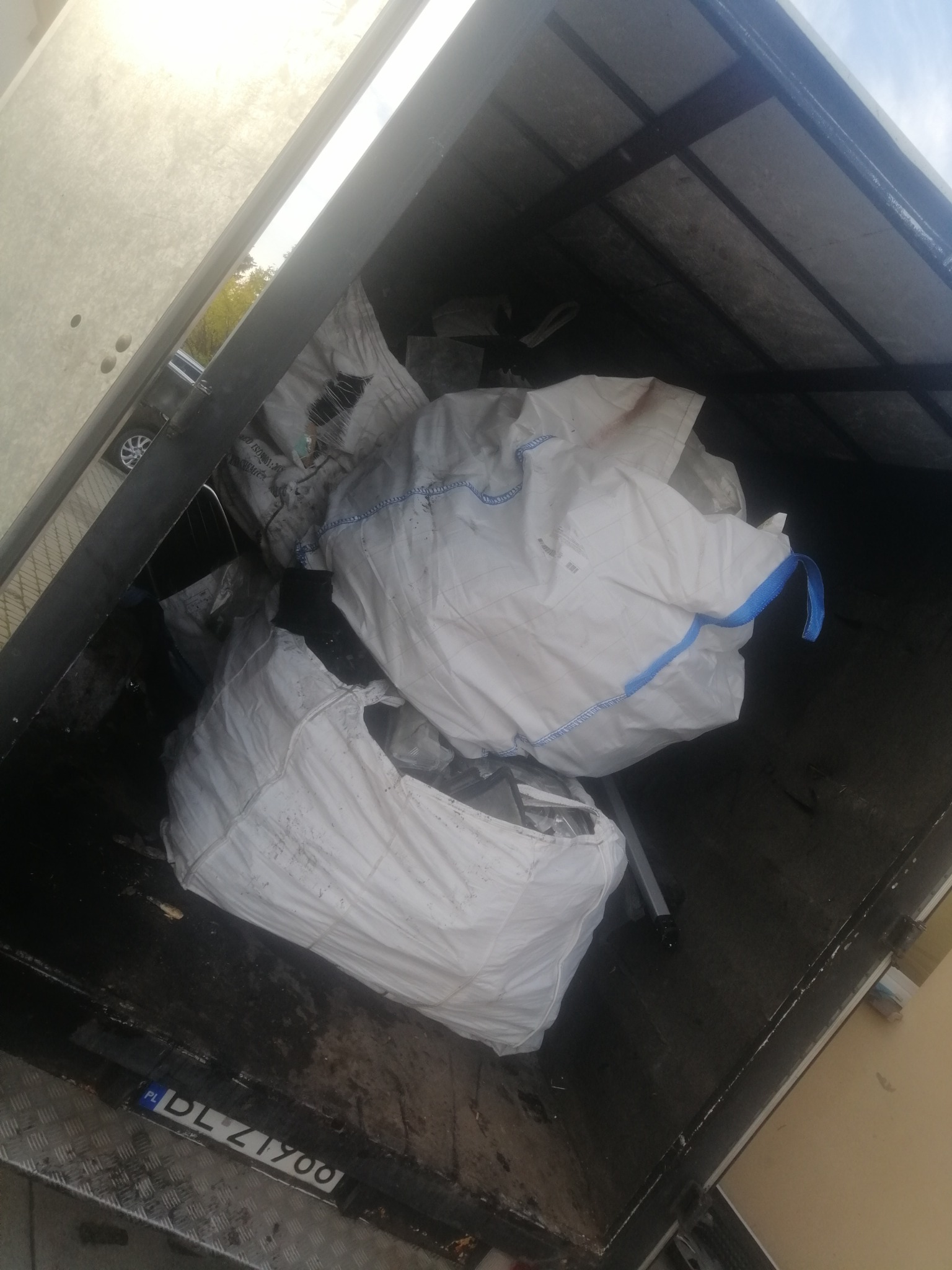 Wnętrze przyczepy z dwoma dużymi, białymi workami typu big bag, wypełnionymi odpadami budowlanymi, widoczne fragmenty gruzu i folii, tablica rejestracyjna pojazdu w dolnej części kadru.