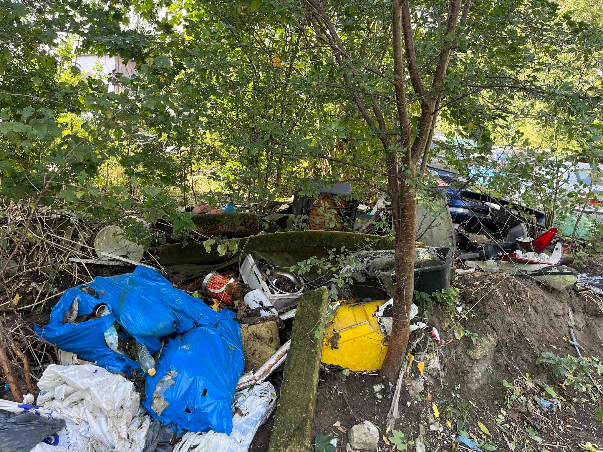 Stos gruzu i odpadów, w tym worki, meble i części samochodowe, porzucony w zaroślach.