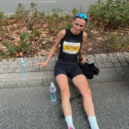 Uśmiechnięta biegaczka w czarnym stroju, numer 22160, siedzi po biegu na chodniku z butelkami wody. W tle banery Magazyn Bieganie.pl i ACCREO.