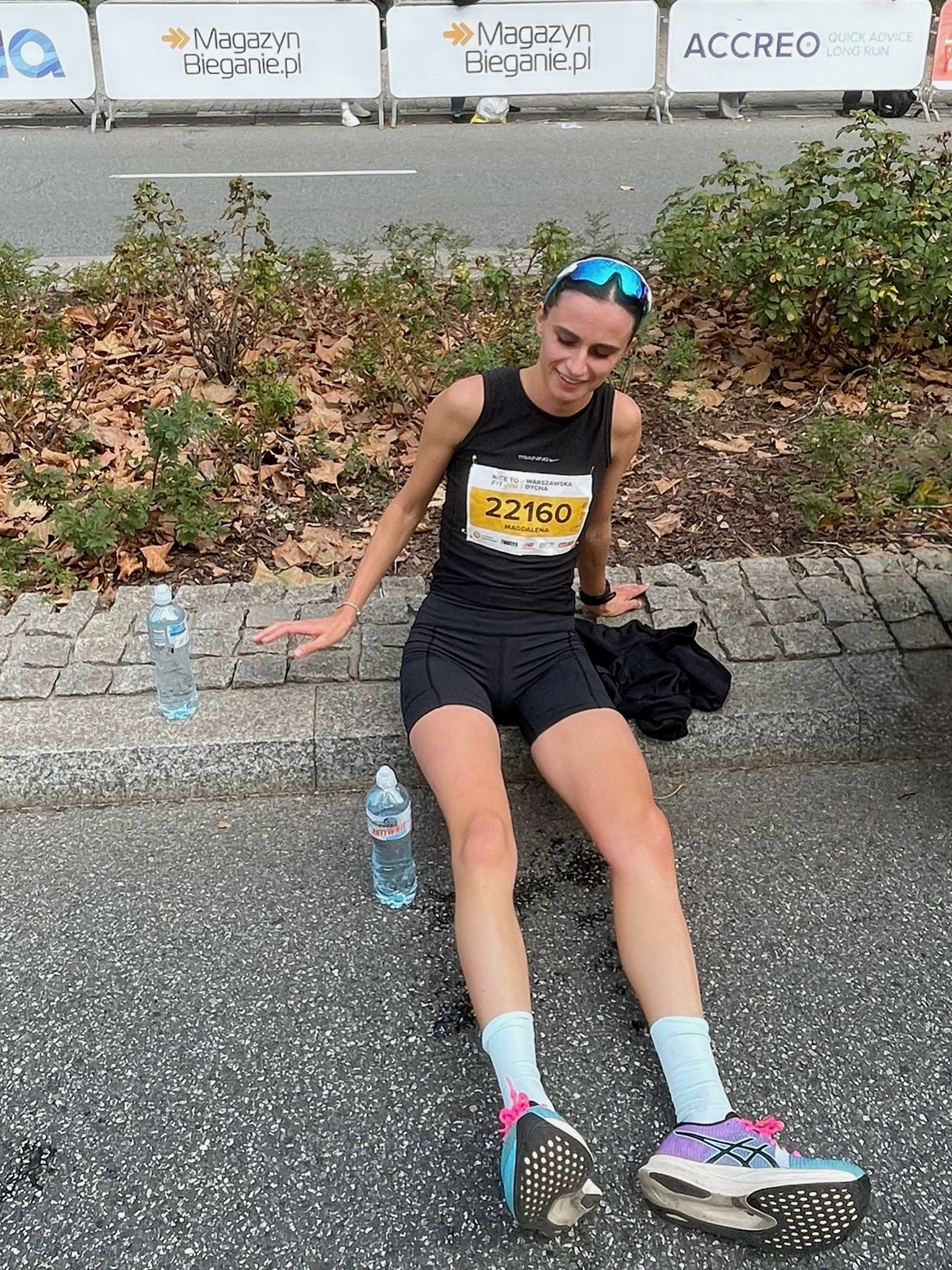 Uśmiechnięta biegaczka w czarnym stroju, numer 22160, siedzi po biegu na chodniku z butelkami wody. W tle banery Magazyn Bieganie.pl i ACCREO.