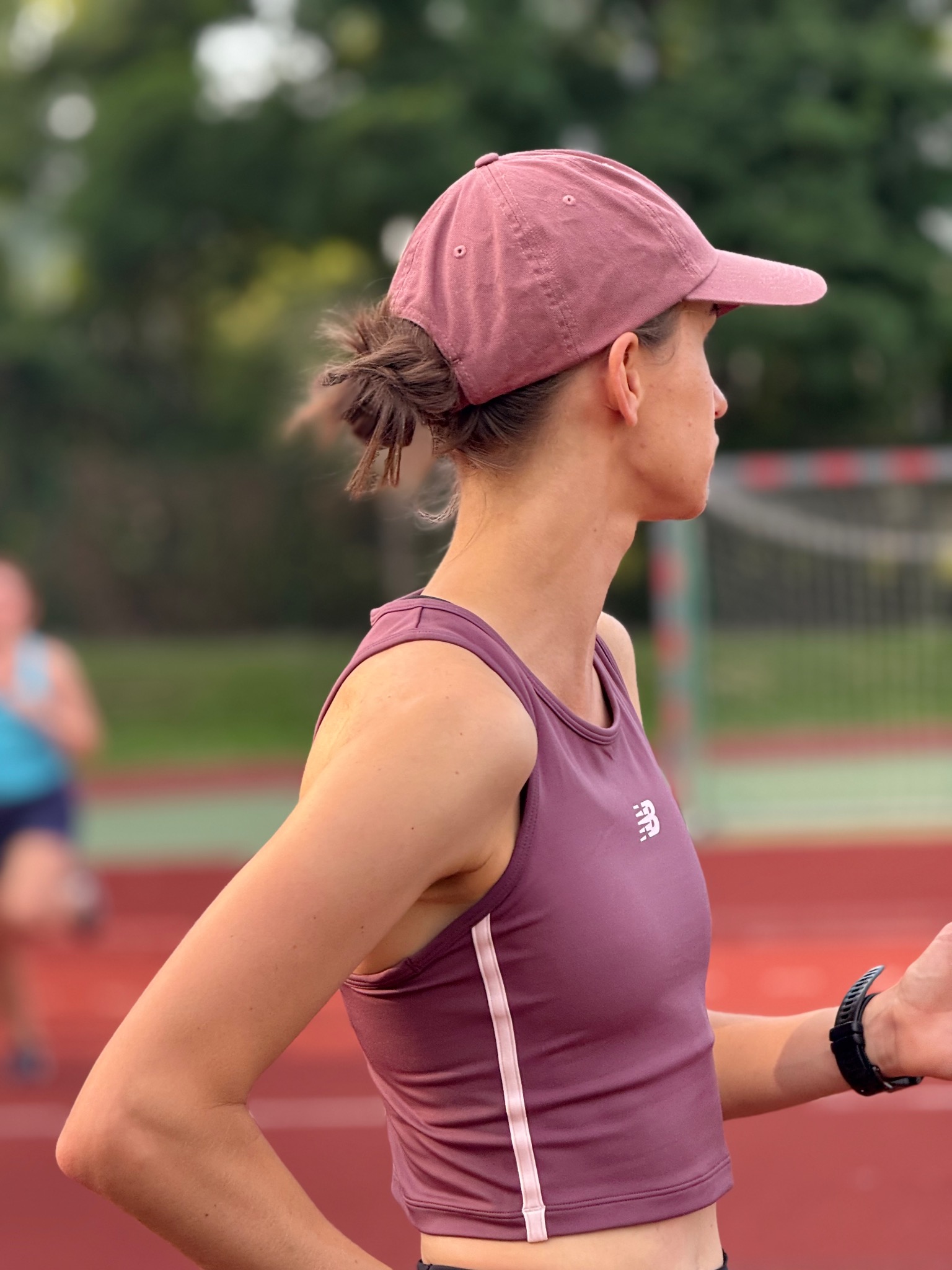 Kobieta w sportowym stroju i czapce patrzy w dal na bieżni. Widoczny zegarek sportowy i sylwetka biegacza w tle. Ujęcie z boku, skupione na detalu stroju i wyrazie twarzy.