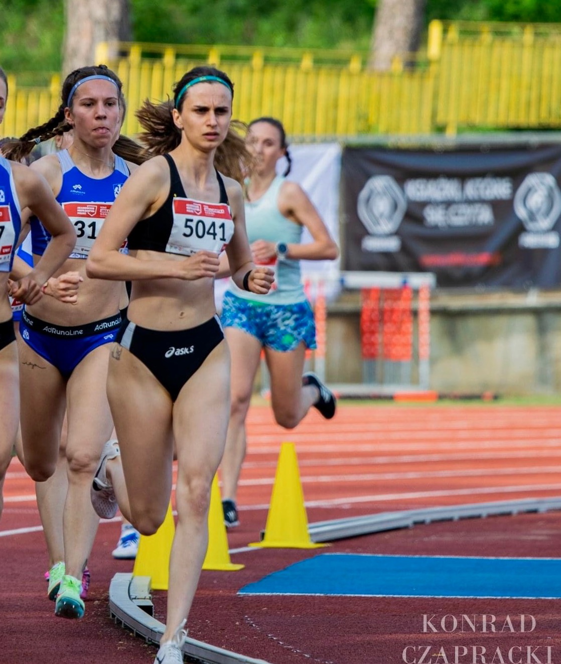 Kobieta z numerem 5041 biegnie w zawodach lekkoatletycznych, widoczny fragment bieżni i inne zawodniczki.