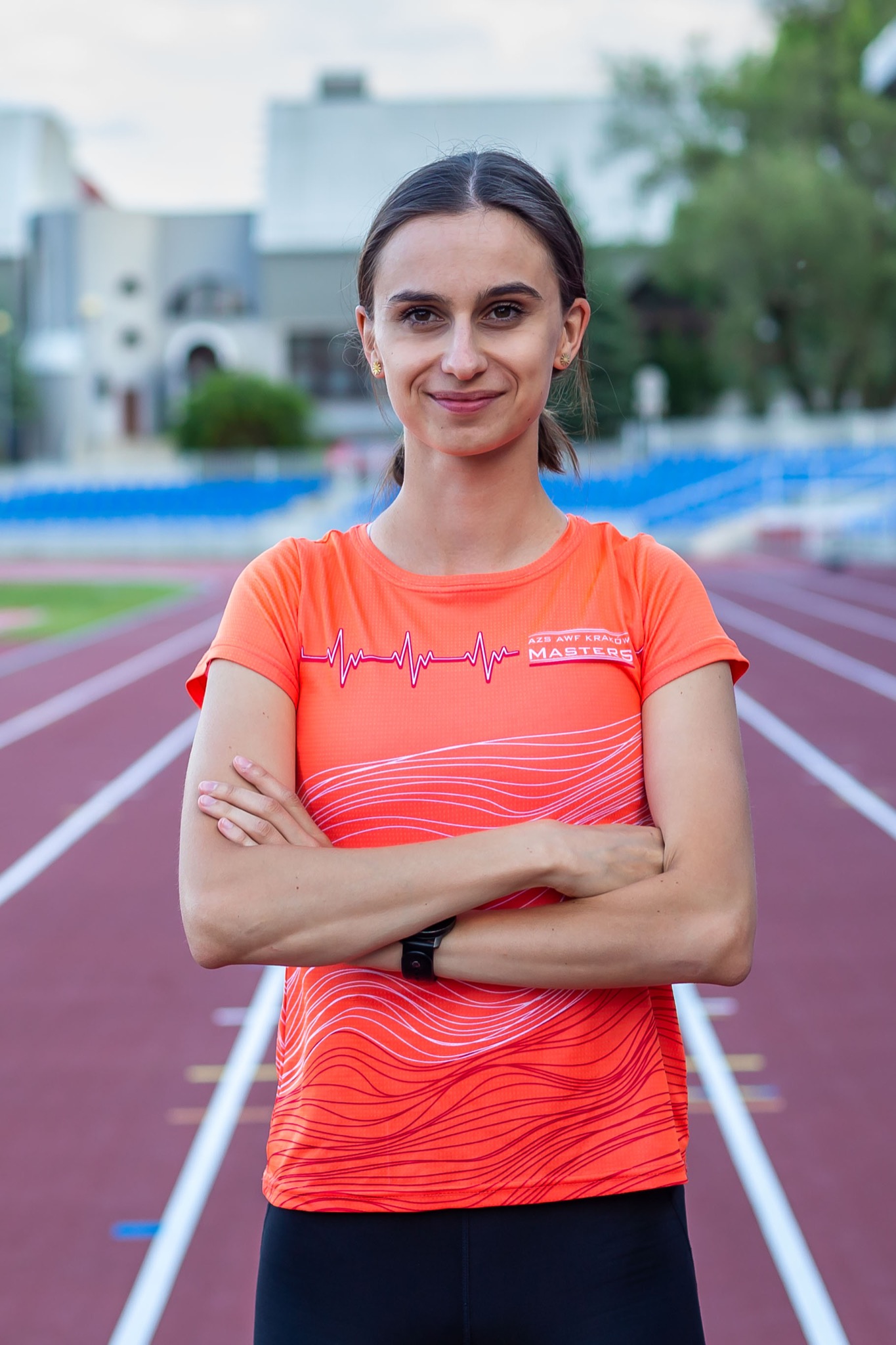 Uśmiechnięta kobieta w pomarańczowej koszulce z nadrukiem EKG i napisem AZS AWF Kraków Masters, stojąca na bieżni lekkoatletycznej ze skrzyżowanymi ramionami.