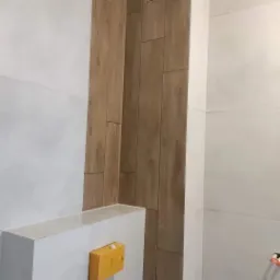 Fragment łazienki z wykończoną ścianą w płytkach imitujących drewno, obok białe płytki i zabudowa stelaża WC z żółtą skrzynką narzędziową.