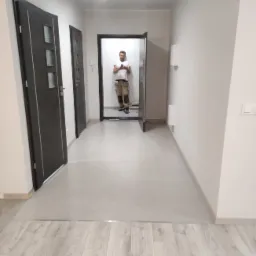 Korytarz po remoncie z szarą podłogą z płytek i paneli podłogowych, widoczne dwie pary drzwi i pracownik w oddali robiący zdjęcie telefonem.