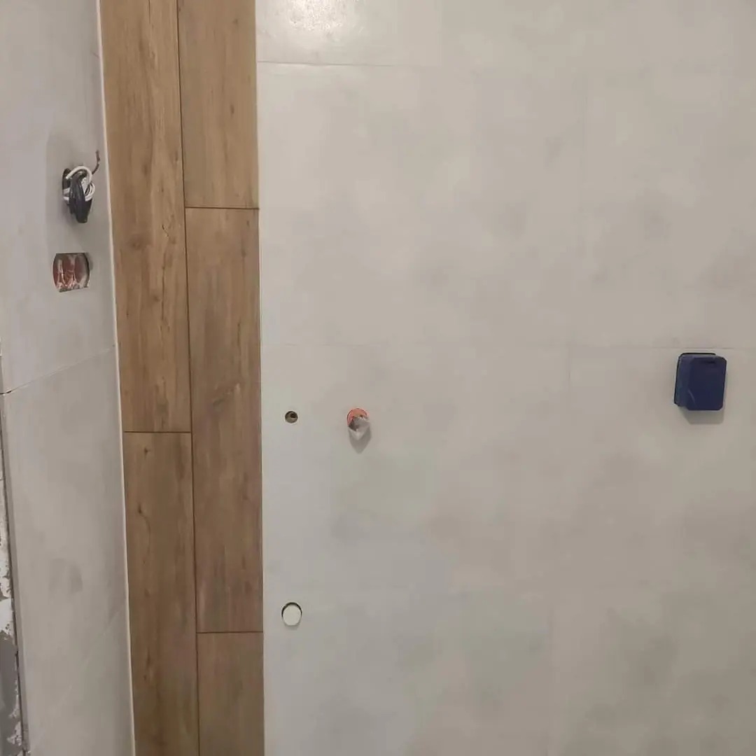 Fragment łazienki w trakcie remontu: ściana z przygotowaną instalacją hydrauliczną i elektryczną, częściowo wyłożona płytkami imitującymi drewno, widoczne niedoskonałości w postaci otworów...