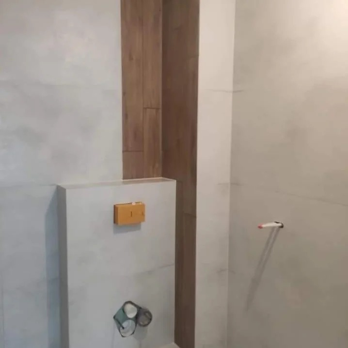 Ściana łazienki wykończona szarą, matową płytką z drewnianym panelem dekoracyjnym, widoczne elementy instalacji wodnej i przycisk spłuczki.