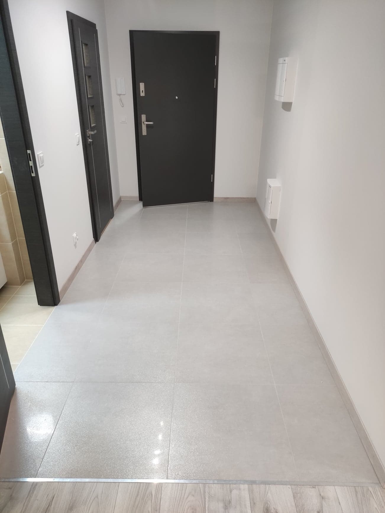 Jasne, kwadratowe płytki podłogowe w korytarzu, widoczne dwie pary drzwi i skrzynki elektryczne na ścianie.