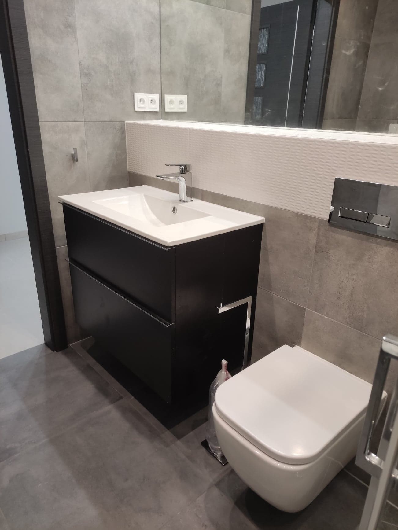 Nowoczesna łazienka z szarymi płytkami, czarną szafką pod umywalkę z chromowanymi uchwytami, podwieszaną toaletą i dużym lustrem z białym, teksturowanym pasem dekoracyjnym.