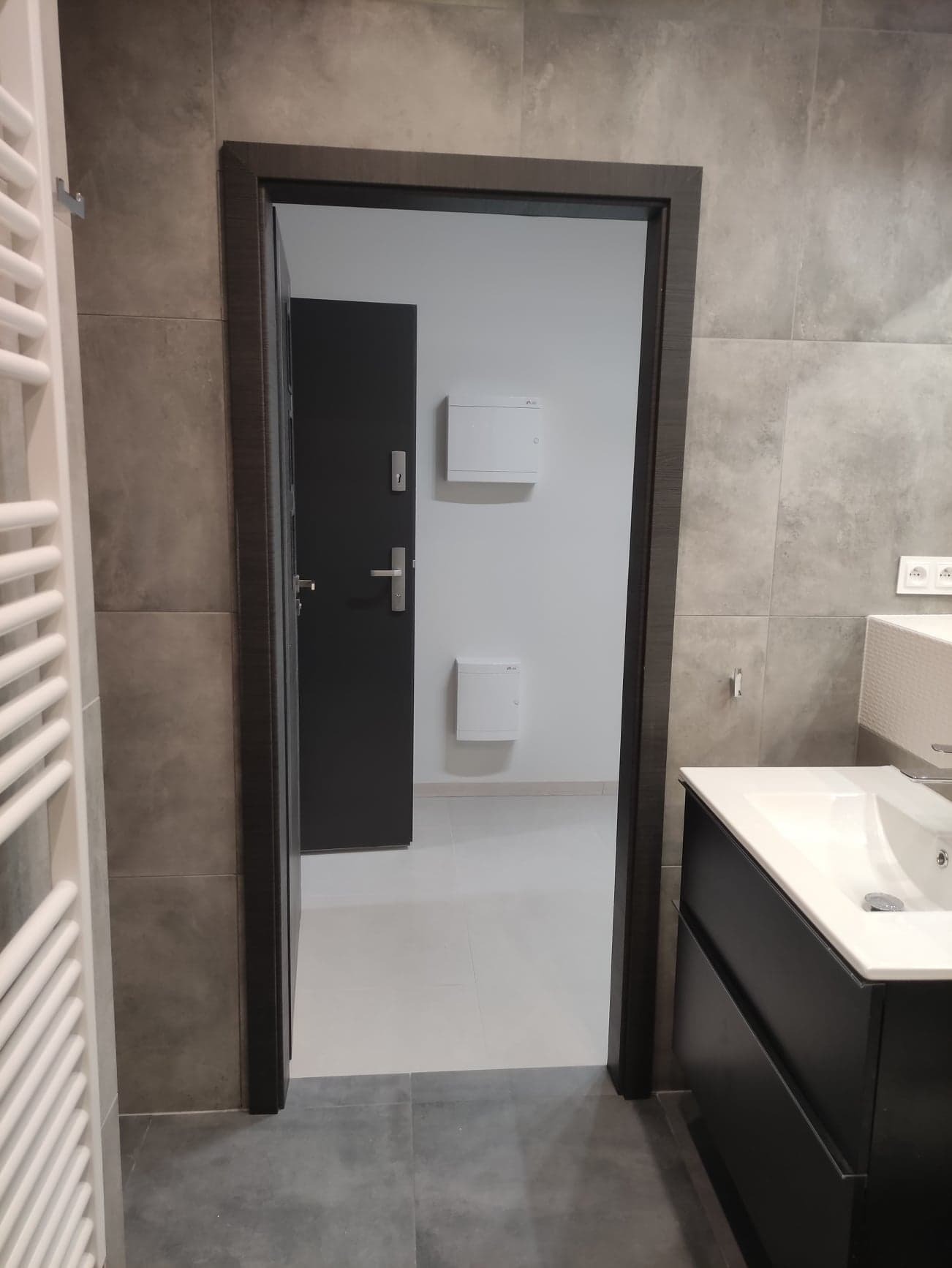 Nowoczesna łazienka z szarymi płytkami, widok na korytarz przez otwarte drzwi, biała umywalka z czarną szafką i grzejnik drabinkowy po lewej stronie.