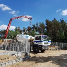 Dwa samochody betonomieszarki na placu budowy, jeden z wysięgnikiem pompy do betonu, w tle las i błękitne niebo z chmurami.