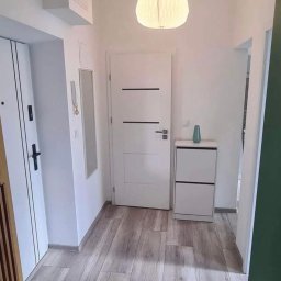 Osw Sens - Nowoczesny przedpokój z białą szafką na buty, drzwiami wewnętrznymi z poziomymi czarnymi paskami i panelami podłogowymi imitującymi drewno ułożonymi wzdłuż korytarza.