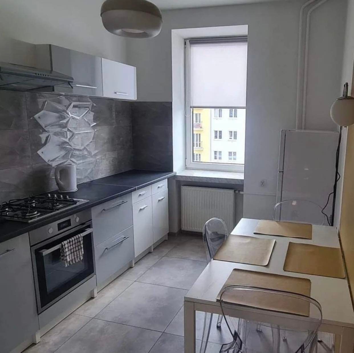 Jasna kuchnia z szarymi płytkami o geometrycznym wzorze, białymi szafkami, czarnym blatem, kuchenką gazową i piekarnikiem, oraz białym stołem z transparentnymi krzesłami.