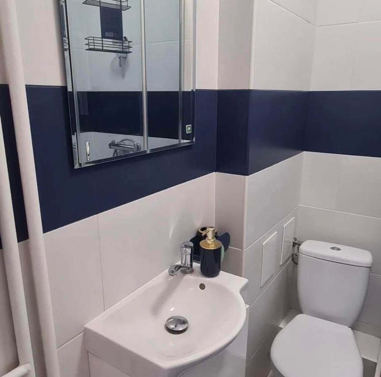 Nowoczesna łazienka z białymi i granatowymi płytkami, umywalką z chromowaną baterią, toaletą i lustrem szafkowym.
