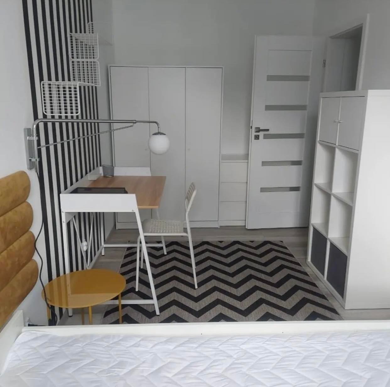 Pokój z biurkiem, krzesłem, szafą i tapetą w czarno-białe pasy. Na ścianie metalowe koszyki. Na podłodze dywan w zygzaki.