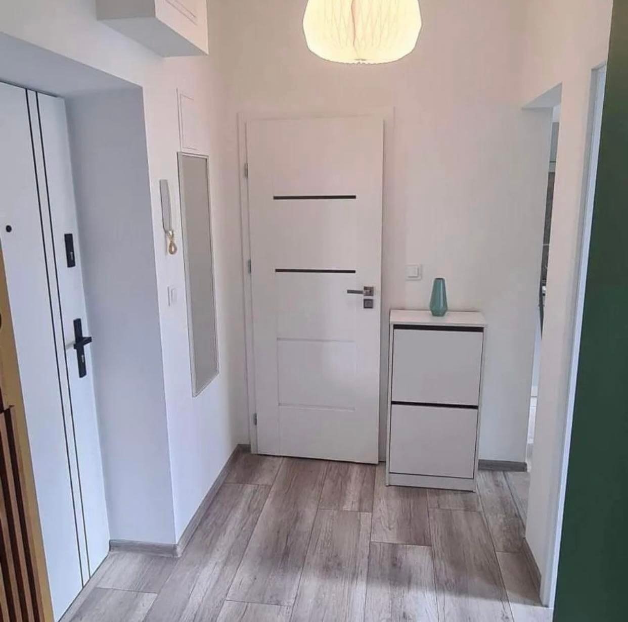 Nowoczesny przedpokój z białą szafką na buty, drzwiami wewnętrznymi z poziomymi czarnymi paskami i panelami podłogowymi imitującymi drewno ułożonymi wzdłuż korytarza.