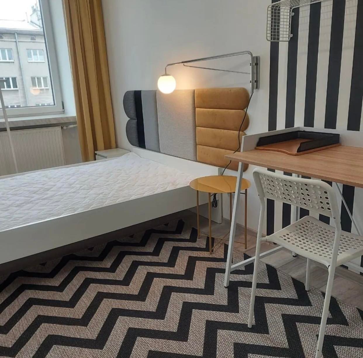 Nowoczesna sypialnia z łóżkiem z tapicerowanym zagłówkiem w odcieniach szarości i beżu, biurkiem z jasnego drewna i białym krzesłem, dywan w zygzaki, lampa ścienna i czarno-biała tapeta w paski.