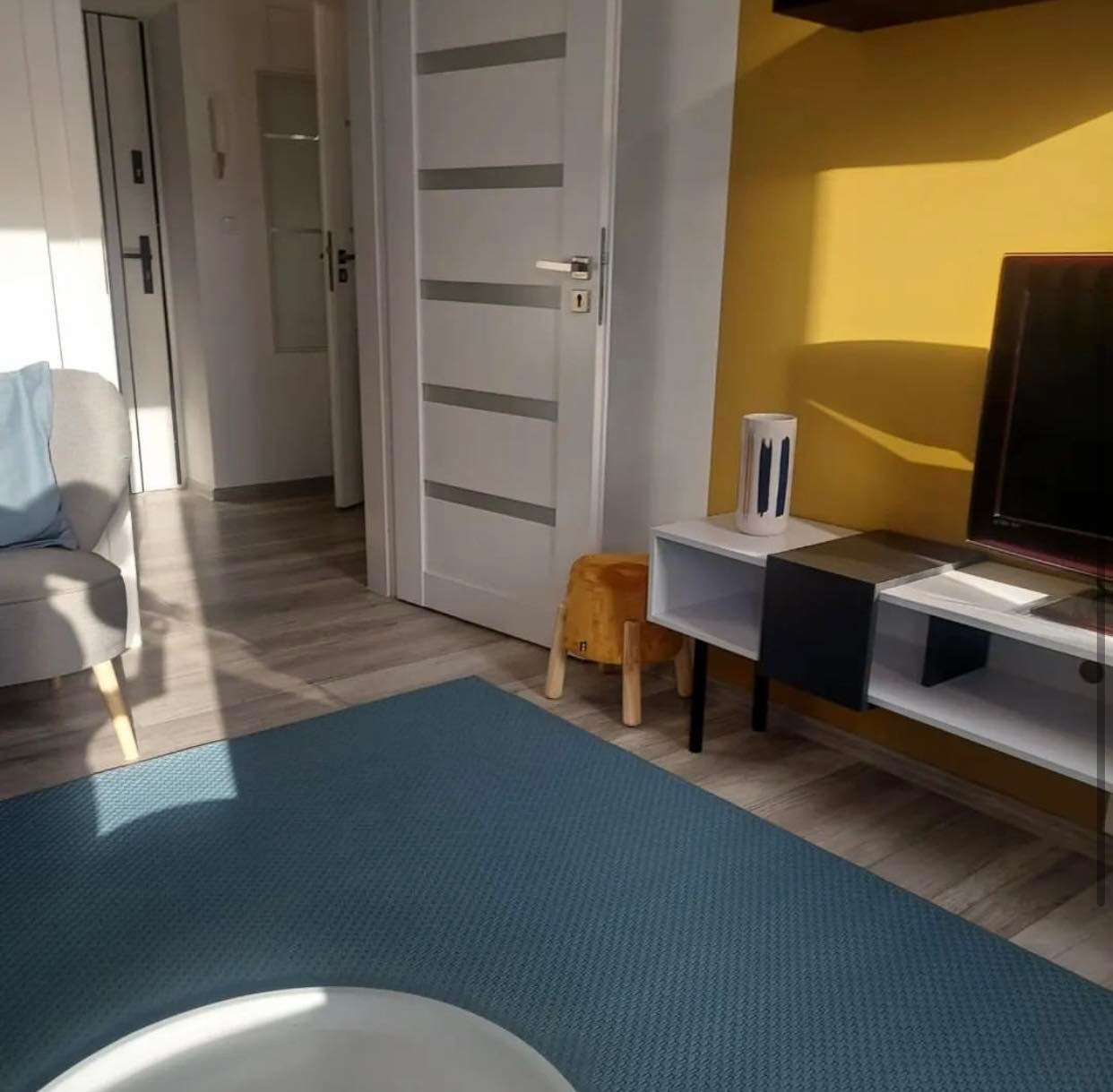 Nowoczesny salon z żółtą ścianą, białą szafką RTV, szarym fotelem i niebieskim dywanem, widoczna biała drzwi z szarymi wstawkami.