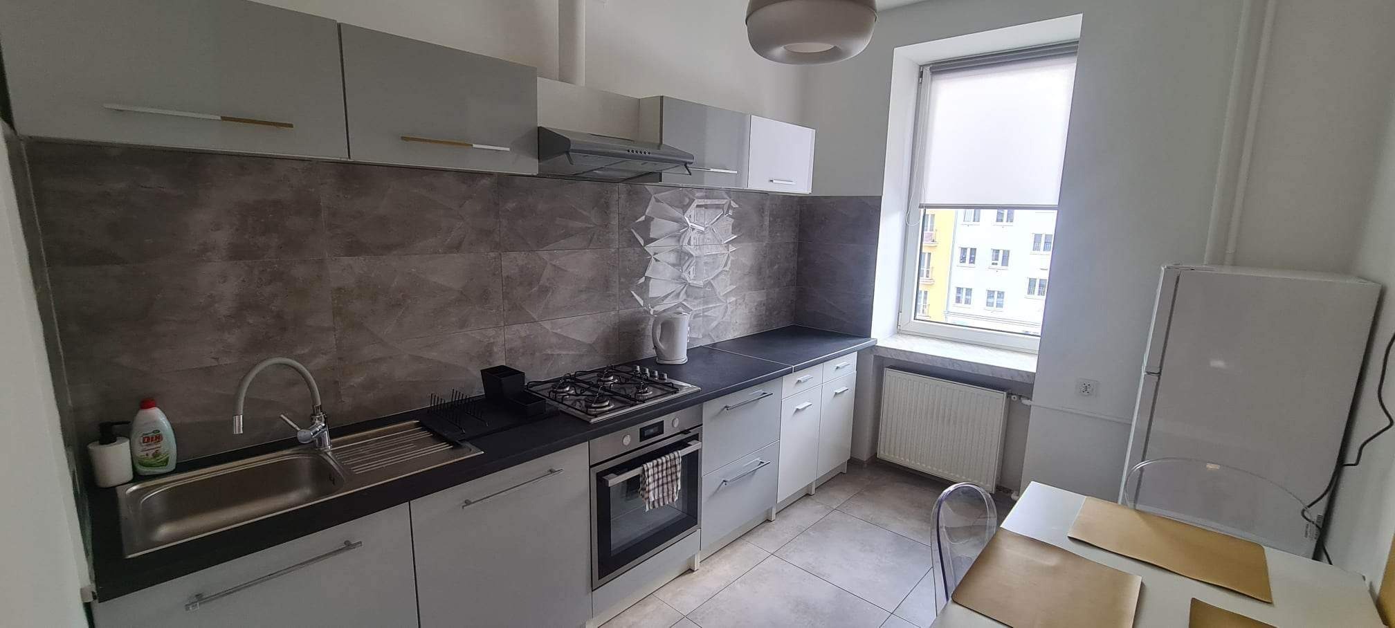 Jasna kuchnia z szarymi szafkami, ciemnym blatem i dekoracyjną mozaiką nad blatem, widok na okno z roletą i zabudowę kuchenną z wyposażeniem.