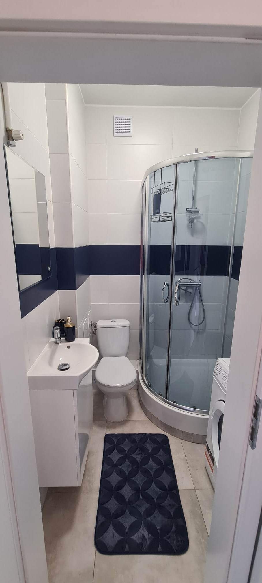 Wykończona łazienka w Warszawie z biało-granatowymi płytkami, kabiną prysznicową z chromowanymi uchwytami, białą umywalką z szafką i ciemnym dywanikiem z geometrycznym wzorem.