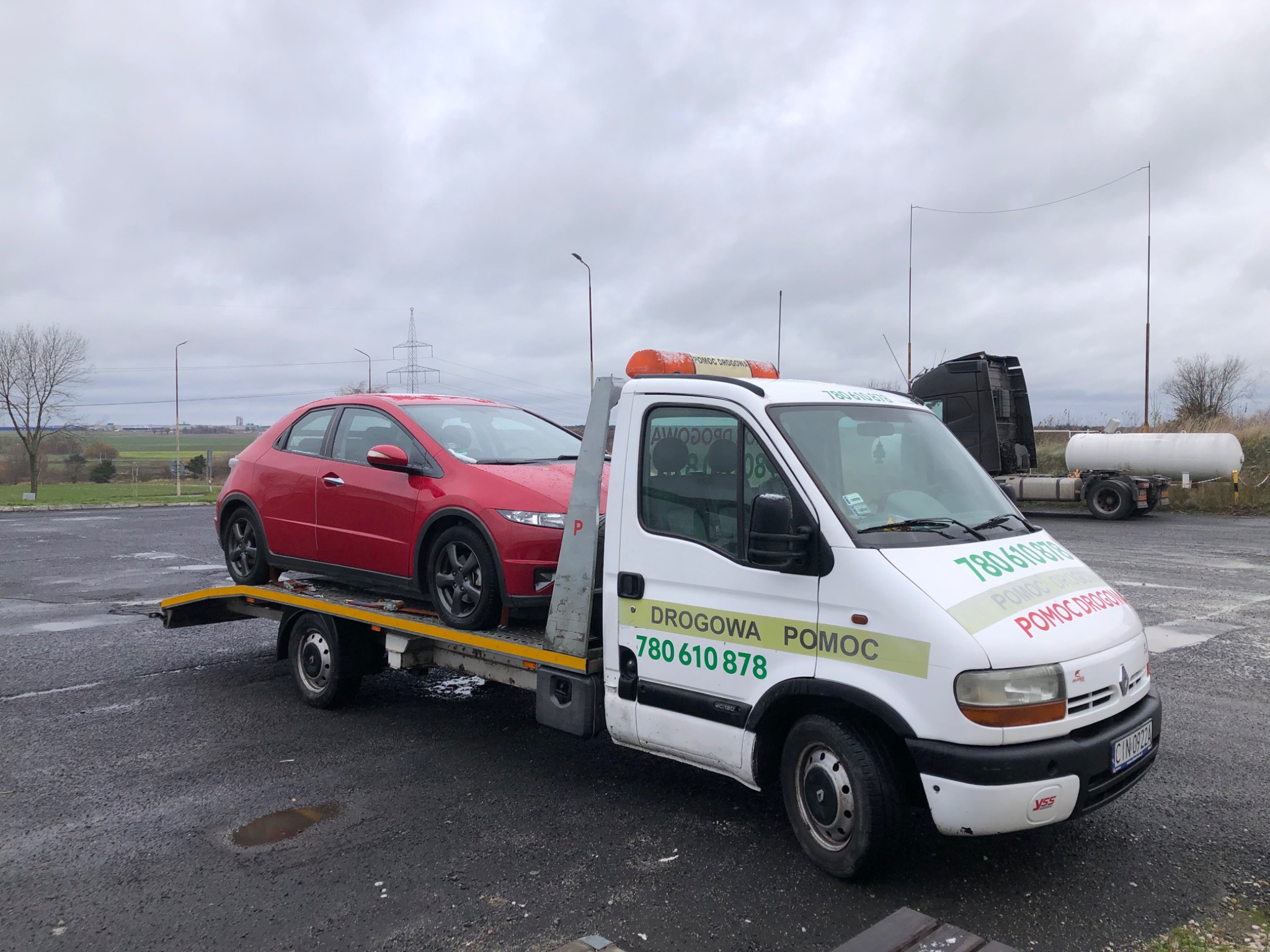 Czerwony samochód osobowy na platformie białej lawety z napisem 'DROGOWA POMOC' na tle szarego parkingu i pochmurnego nieba.