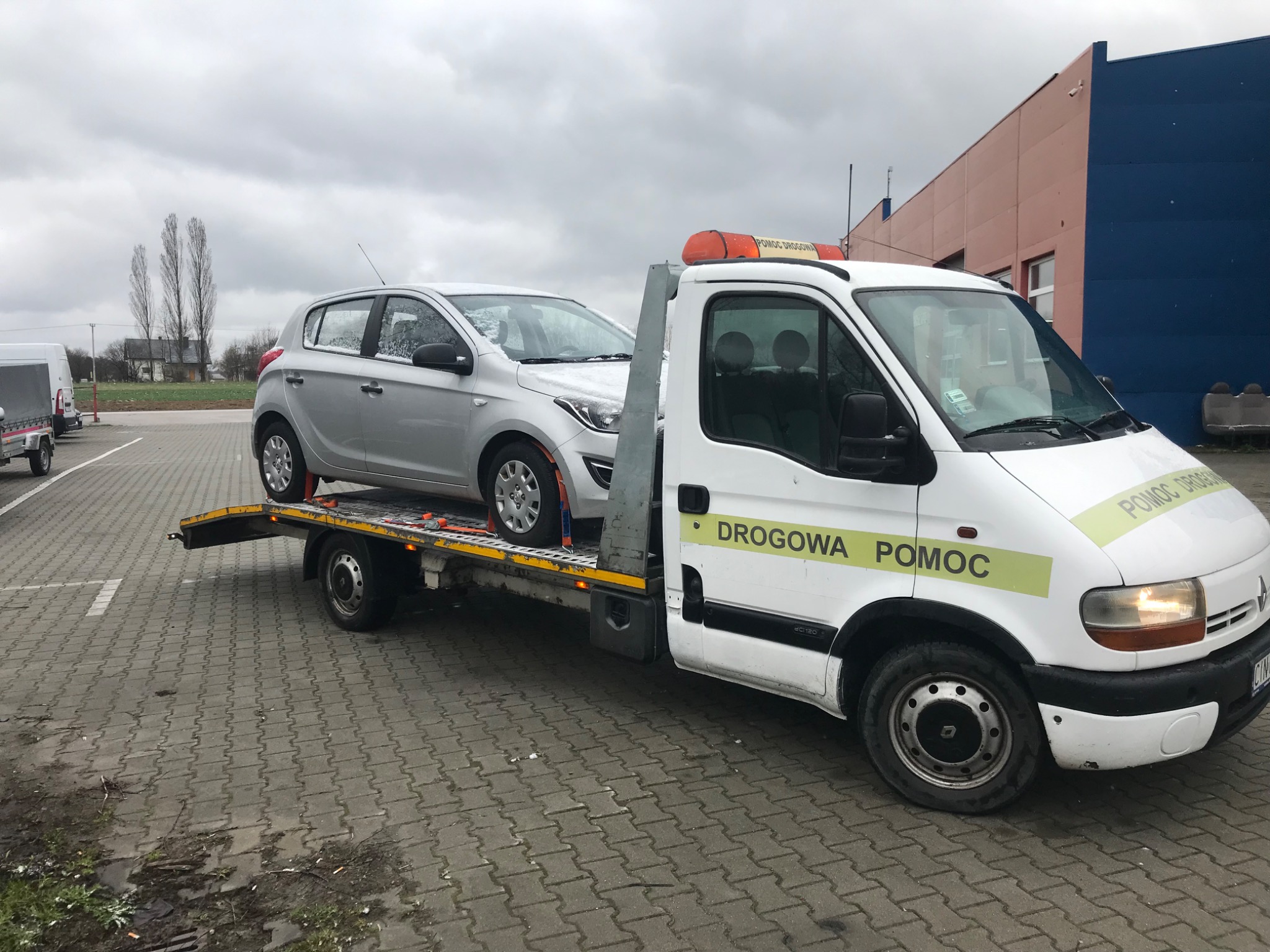 Srebrny samochód osobowy na platformie lawety pomocy drogowej z napisem 'DROGOWA POMOC' na tle szarej kostki brukowej i budynku w pochmurny dzień.