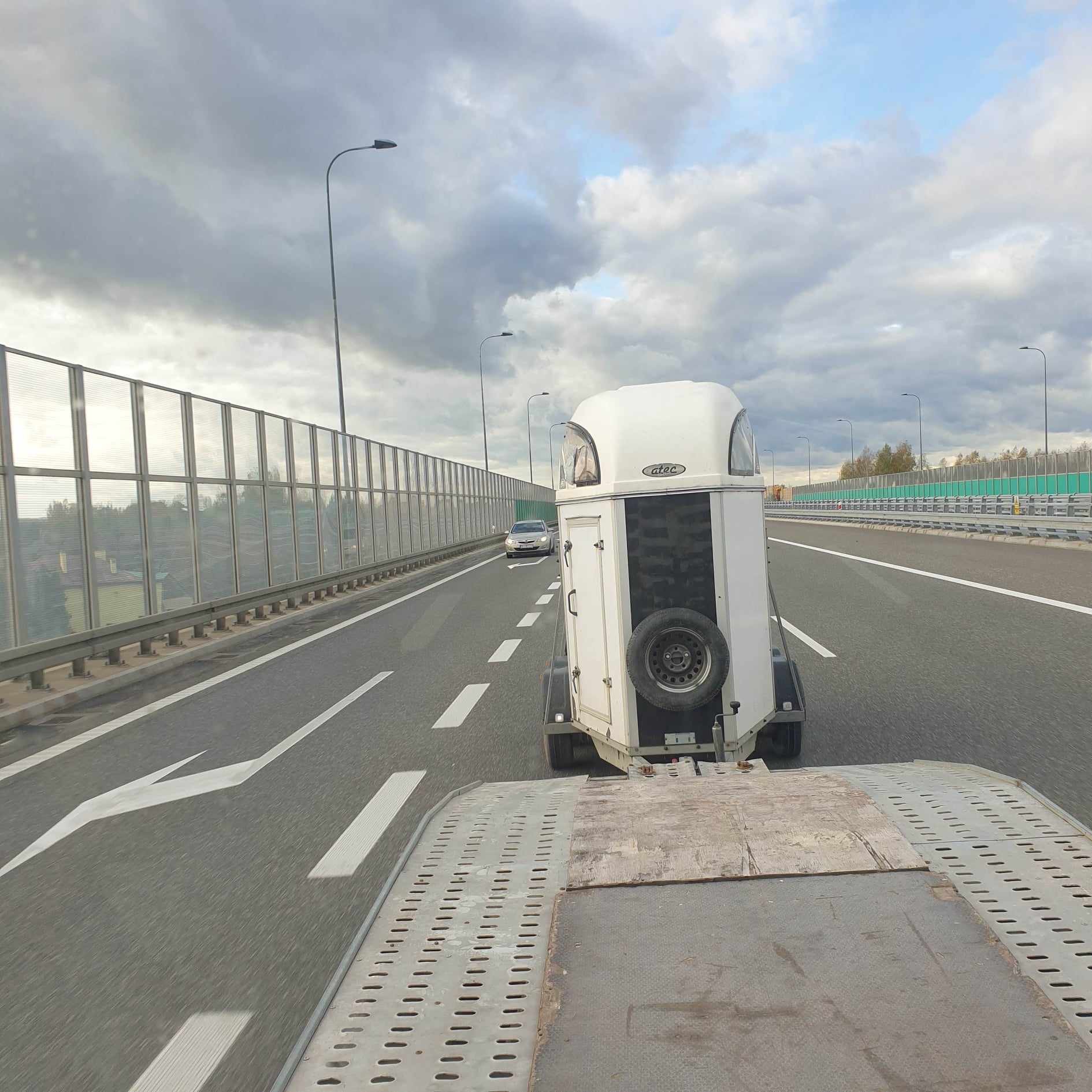 Widok z lawety na białą przyczepę do przewozu koni z kołem zapasowym, holowaną po autostradzie w pochmurny dzień.