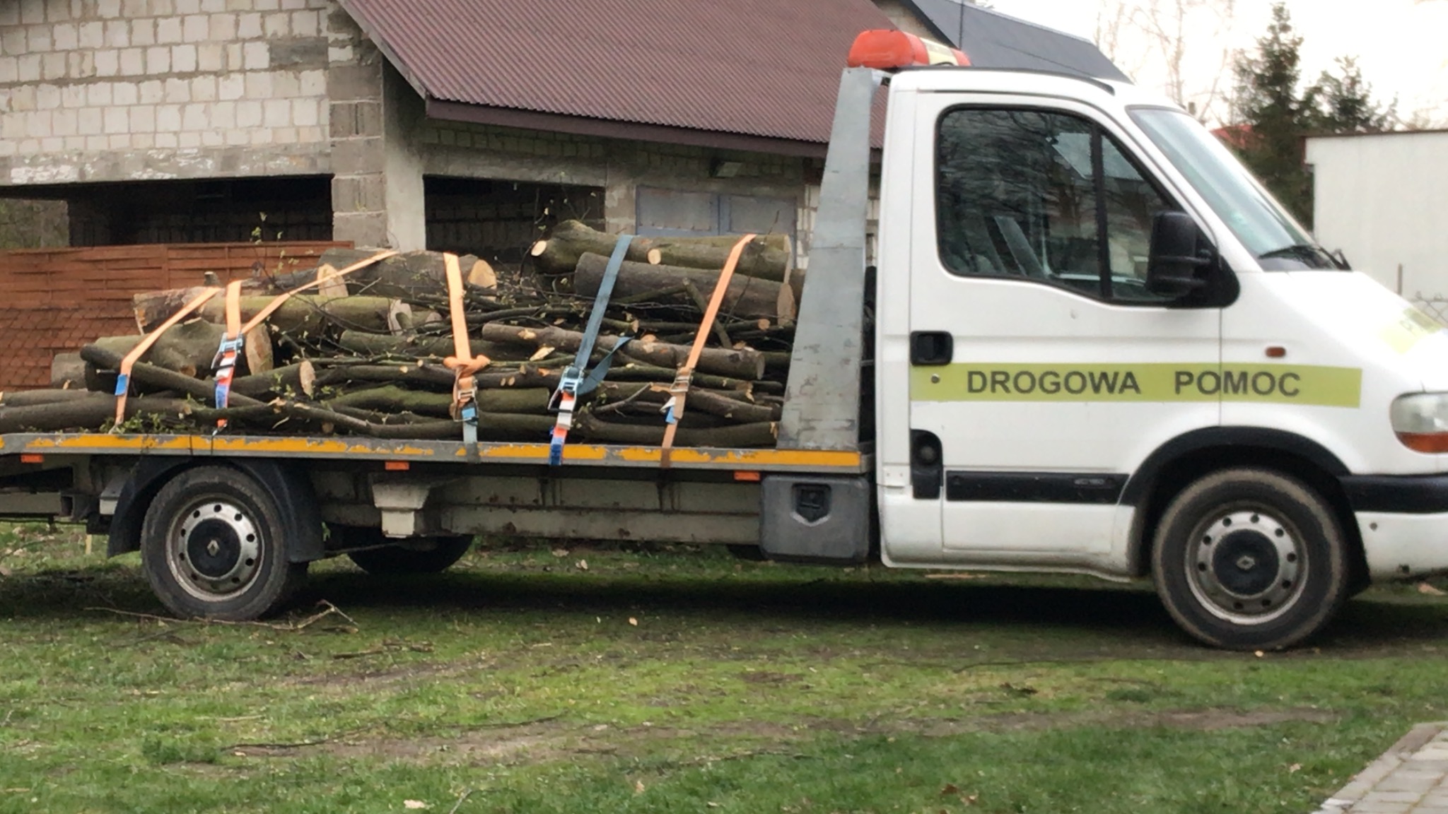 Biała laweta z napisem 'DROGOWA POMOC' przewozi pocięte drewno zabezpieczone pasami transportowymi na tle niedokończonego budynku.