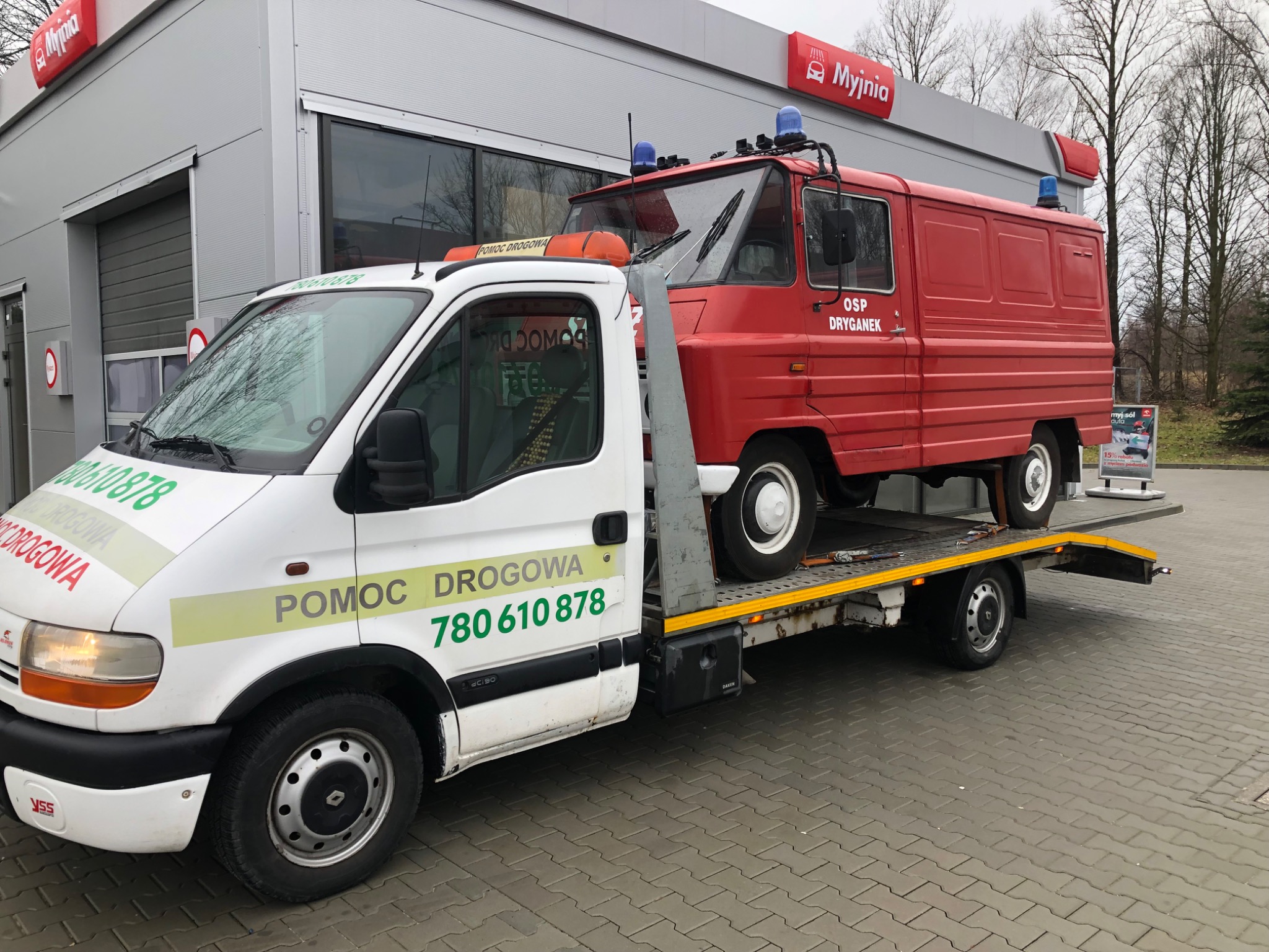 Laweta z białym ciągnikiem i czerwonym wozem strażackim OSP Dryganek na platformie, zaparkowana przed budynkiem myjni samochodowej