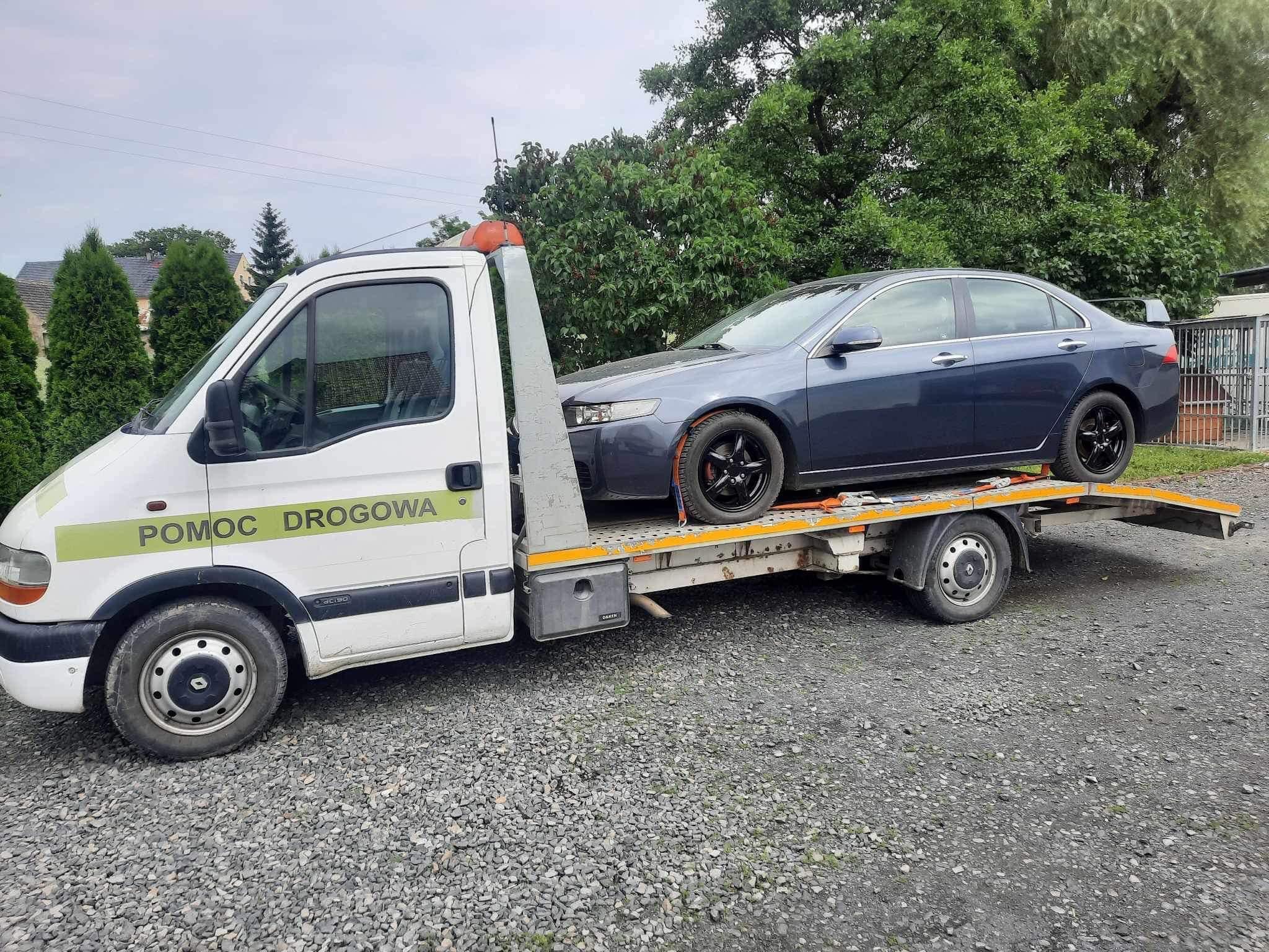 Biała laweta z napisem 'POMOC DROGOWA' przewozi szary samochód osobowy z czarnymi felgami na platformie w pochmurny dzień.