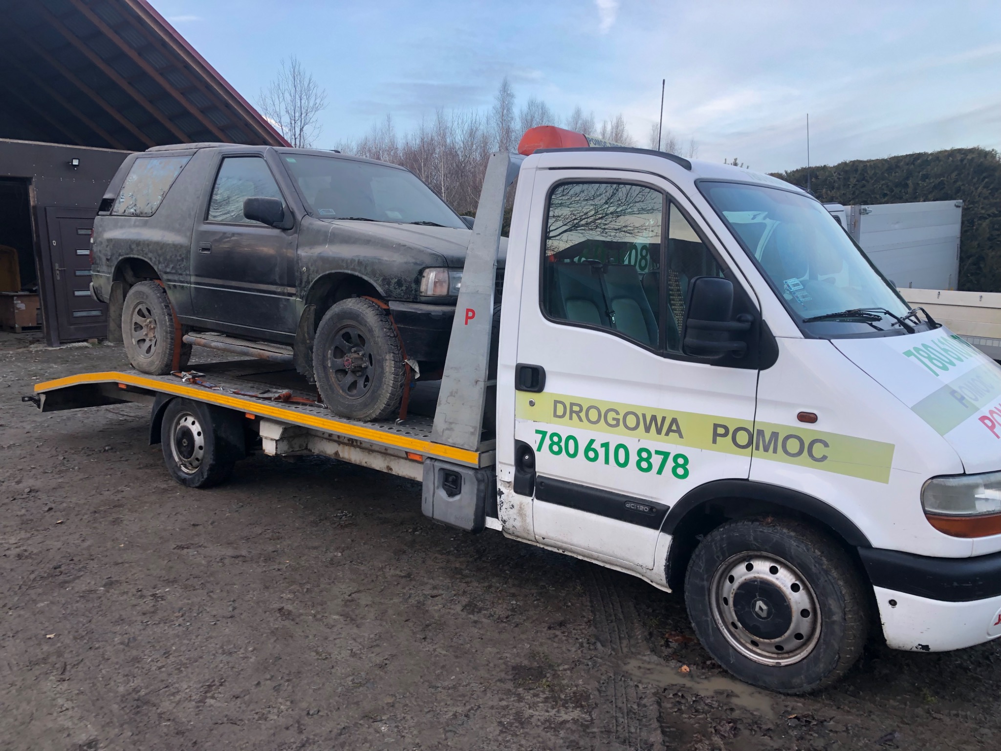 Czarny, lekko zabrudzony samochód terenowy na platformie lawety z napisem 'DROGOWA POMOC' i numerem telefonu, stojącej na błotnistym gruncie.