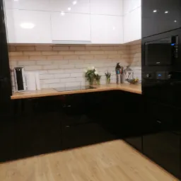 Nowoczesna kuchnia z białymi szafkami górnymi i czarnymi szafkami dolnymi, drewnianym blatem i ceglaną ścianą w tle.