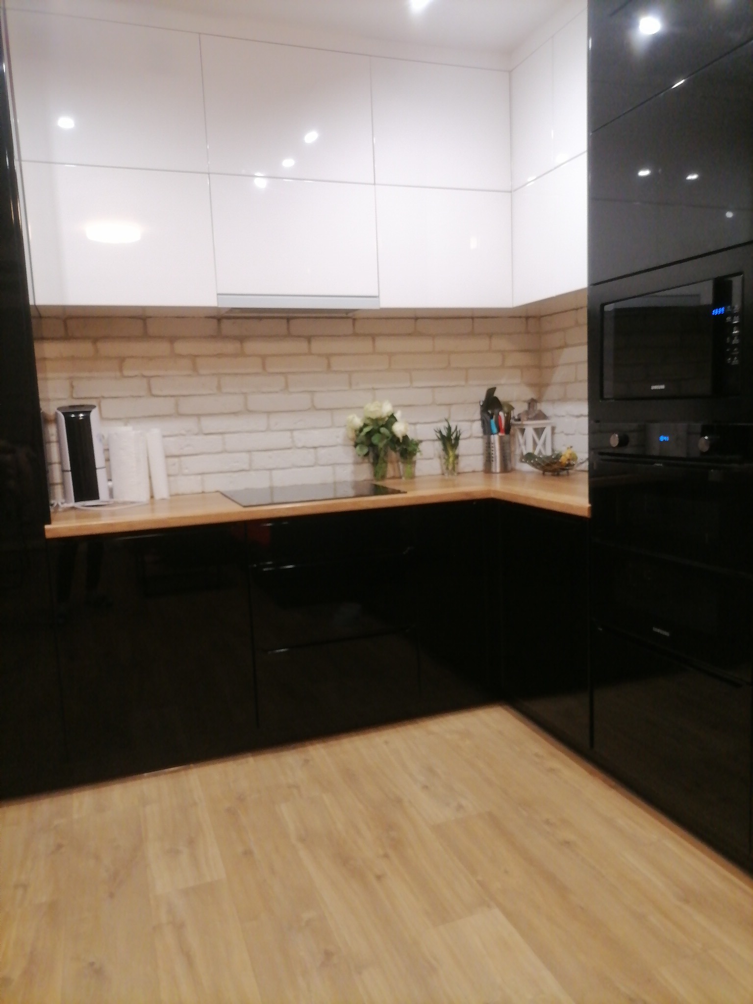 Nowoczesna kuchnia z białymi szafkami górnymi i czarnymi szafkami dolnymi, drewnianym blatem i ceglaną ścianą w tle.