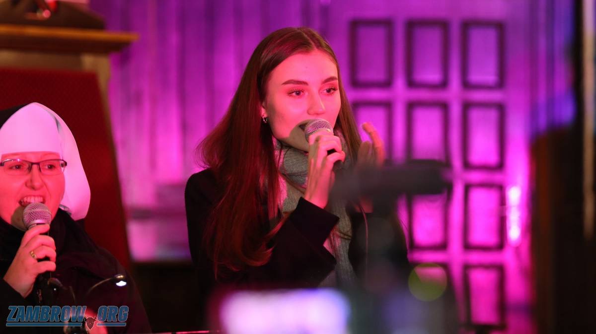 Dwie kobiety śpiewają do mikrofonów na scenie oświetlonej fioletowym światłem, jedna w stroju zakonnym, druga w płaszczu i szaliku.