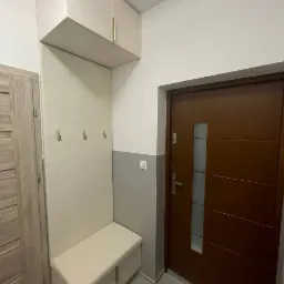 Funkcjonalna zabudowa przedpokoju: szafka górna z złotymi uchwytami, panel z trzema wieszakami i siedzisko z szafką na buty, utrzymane w jasnej kolorystyce, dopasowane do szarej ściany...